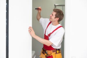 Installation of a lock on the front wooden entrance door. Portrait of young locksmith workman in blue uniform installing door knob. Professional repair service. Maintenance Concept.