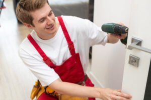 Installation of a lock on the front wooden entrance door. Portrait of young locksmith workman in blue uniform installing door knob. Professional repair service. Maintenance Concept.