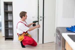 Repair Service Concept. Portrait of smiling handsome young locksmith in blue uniform installing door knob on front wooden entrance door at home or office workplace