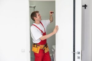 Repair Service Concept. Portrait of smiling handsome young locksmith in blue uniform installing door knob on front wooden entrance door at home or office workplace